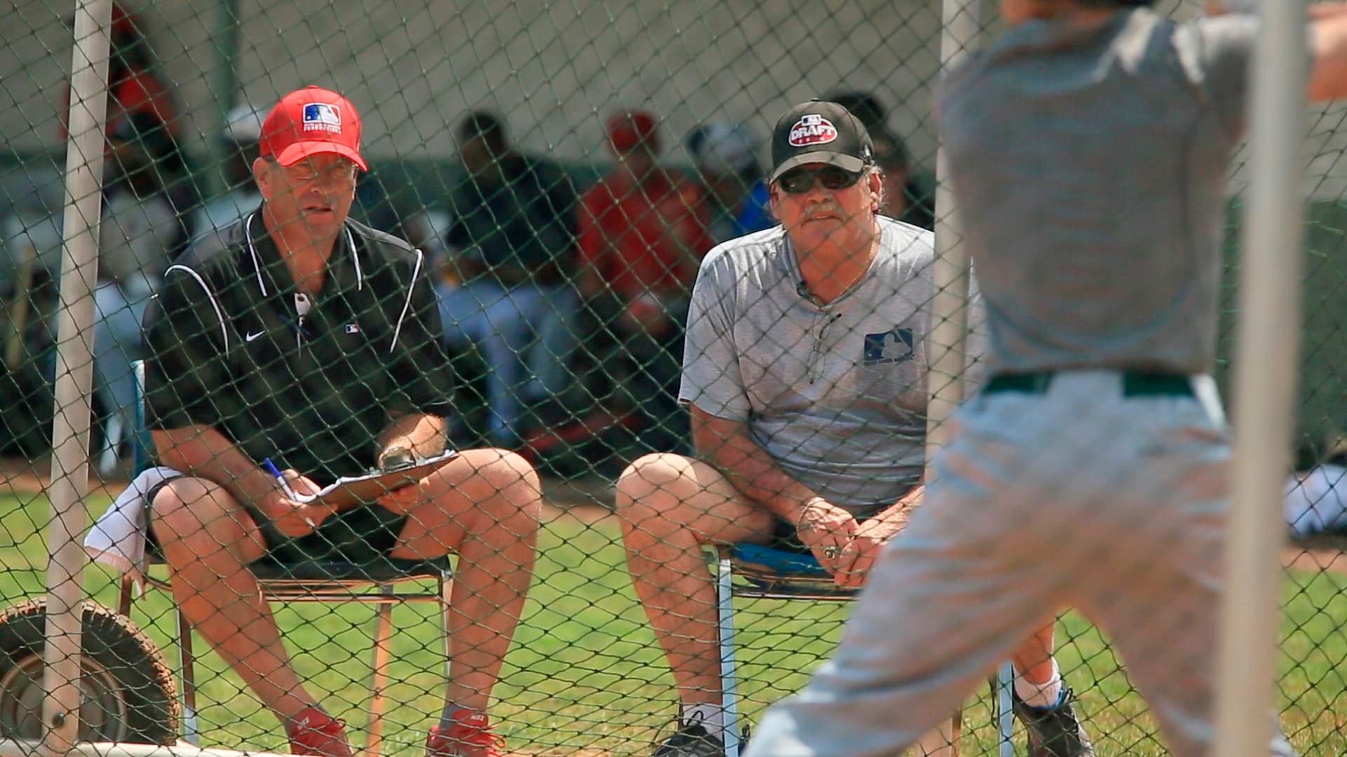 Two baseball scouts sit behind a net, one holding a clipboard, watching a batter practice.