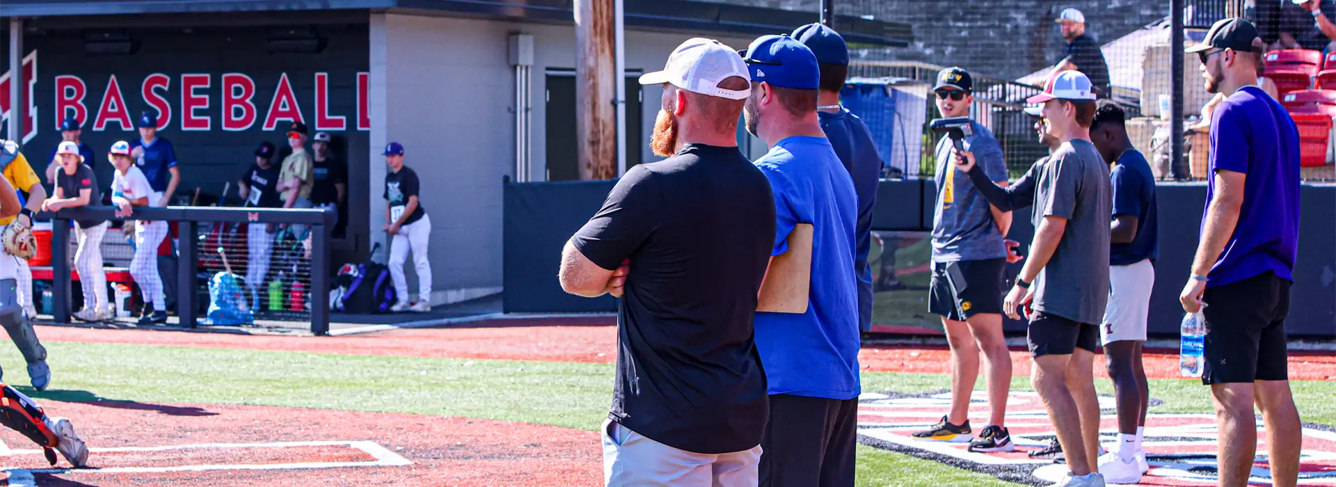 Coaches and scouts monitor a baseball game from the field, one holding a radar gun.