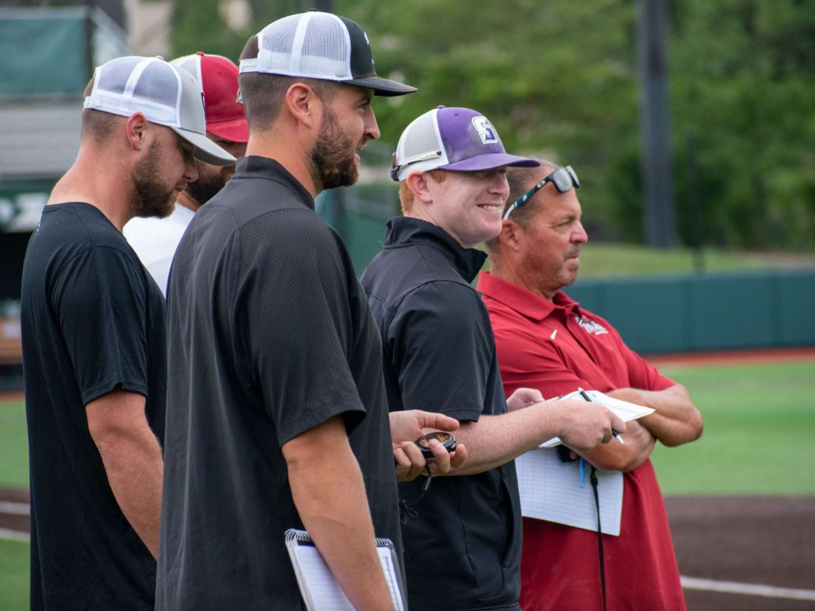 Group of men in baseball caps standing on a field with clipboards and stopwatches.