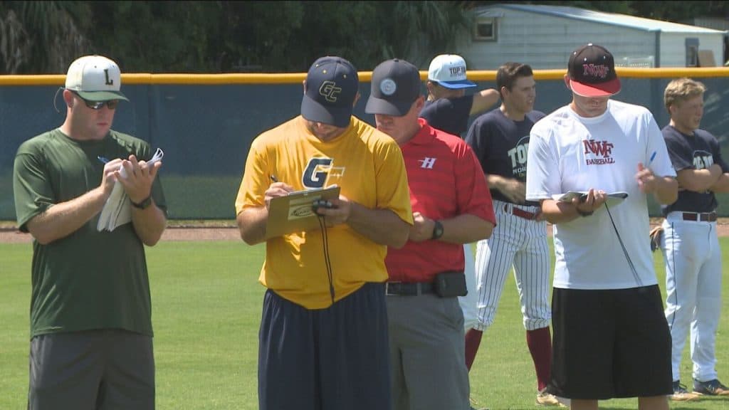 Baseball scouts wearing hats and t-shirts stand on a field taking notes on clipboards.