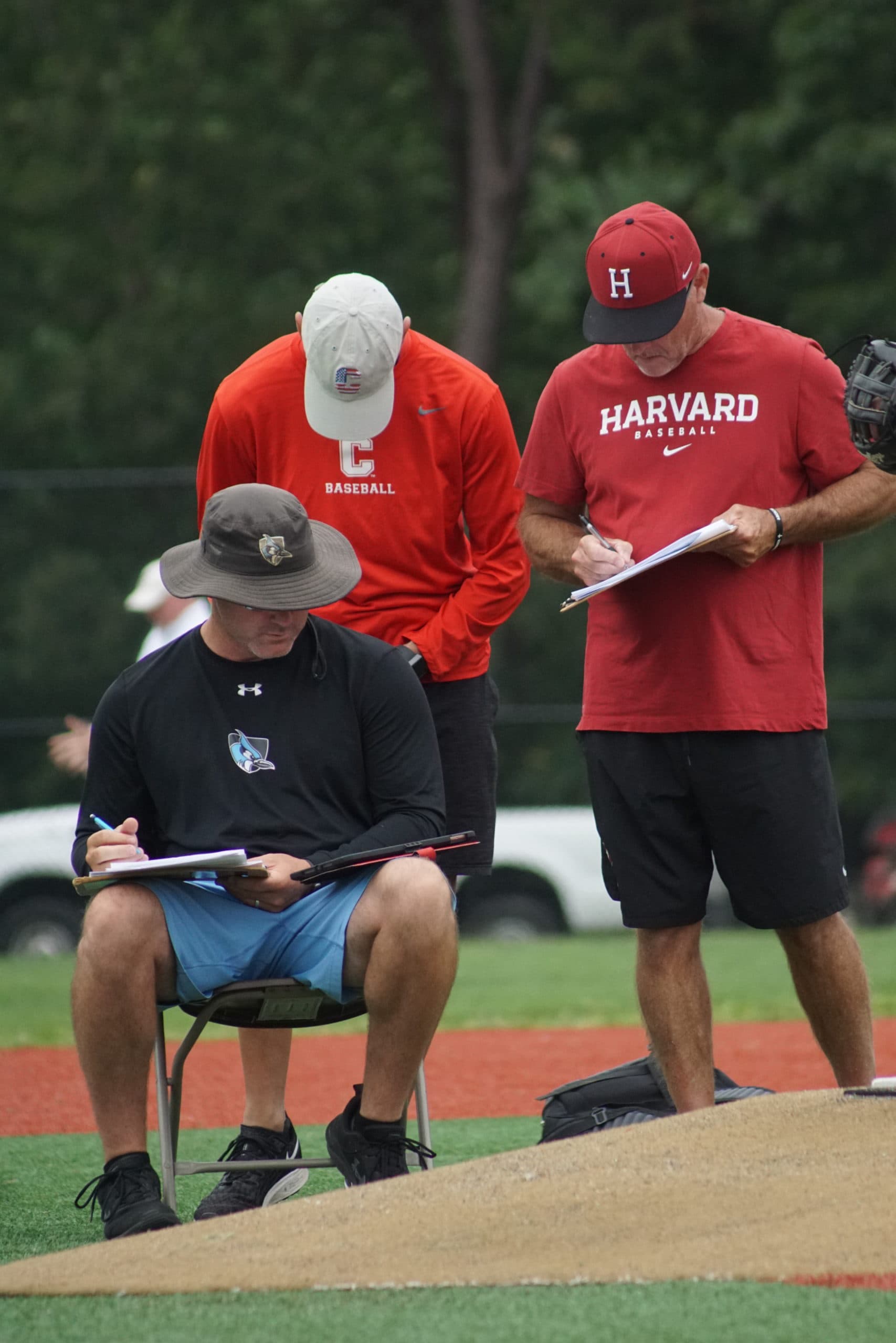 Three baseball coaches in athletic gear take notes on clipboards while observing from the field.